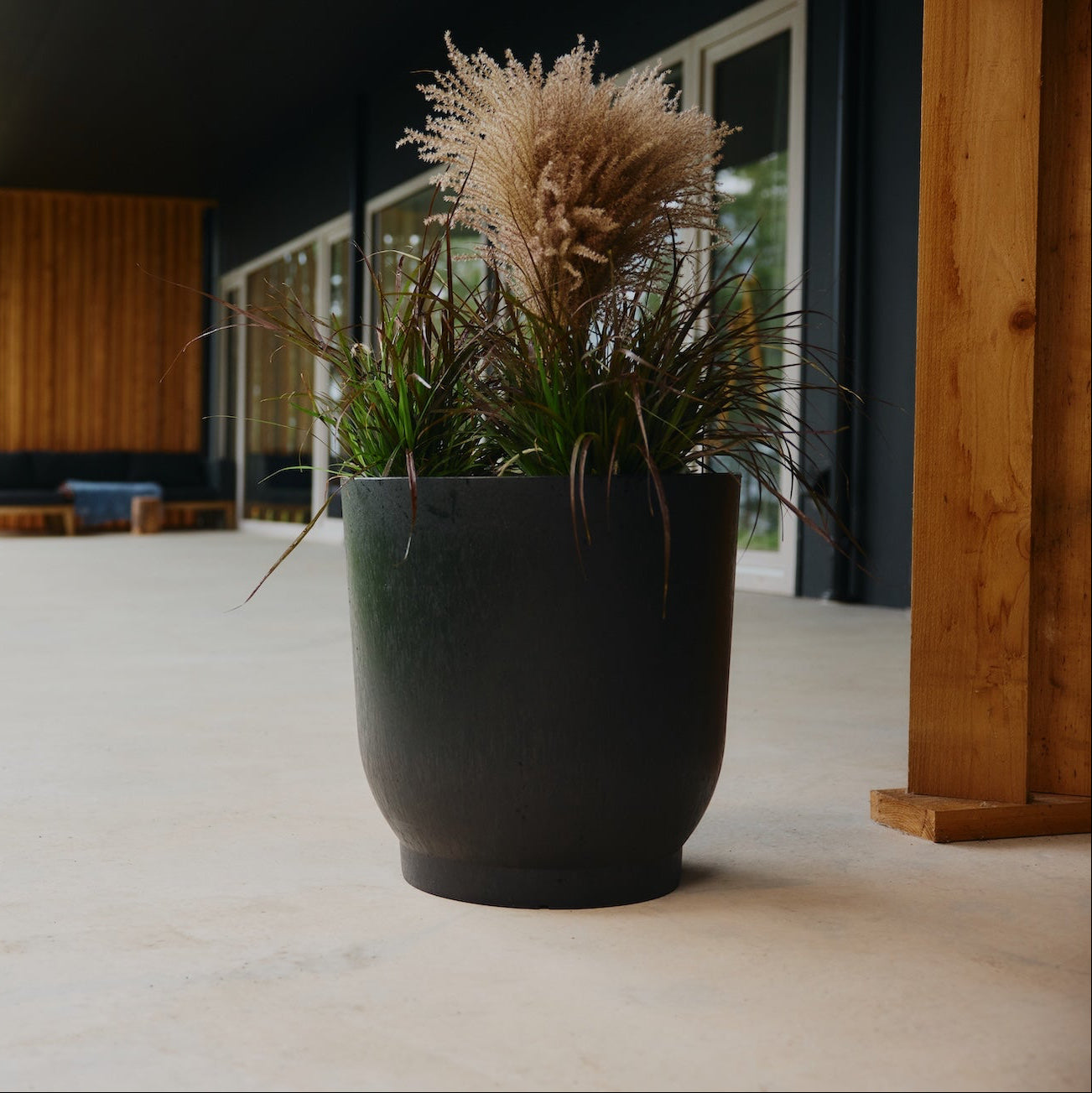 Black planter with dried plants on a patio area with a modern house and greenery in the background.