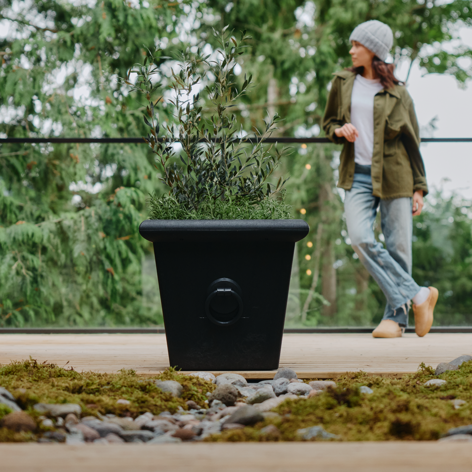 Person walking outdoors near a large black planter with greenery, surrounded by trees and a modern building.