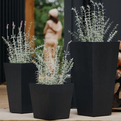 Three black planters with plants on a wooden deck next to stacked firewood.